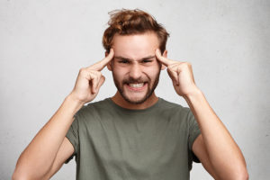 Headshot of young man holds fingers on temples, has bad memory, tries to concentrate and remember details of accident, presses teeth with irritation. Overworked man has headache, feels tired