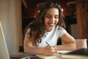 Beautiful student girl with chubby cheeks holding pen handwriting in notebook while learning distantly from home, sitting at table with open laptop, having inspired facial expression, smiling broadly